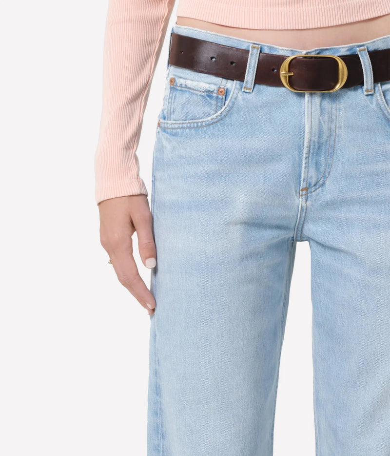 Close-up of a person wearing light-wash blue jeans with a dark brown leather belt featuring a gold-tone buckle, paired with a pale pink ribbed long-sleeve top against a plain light background.