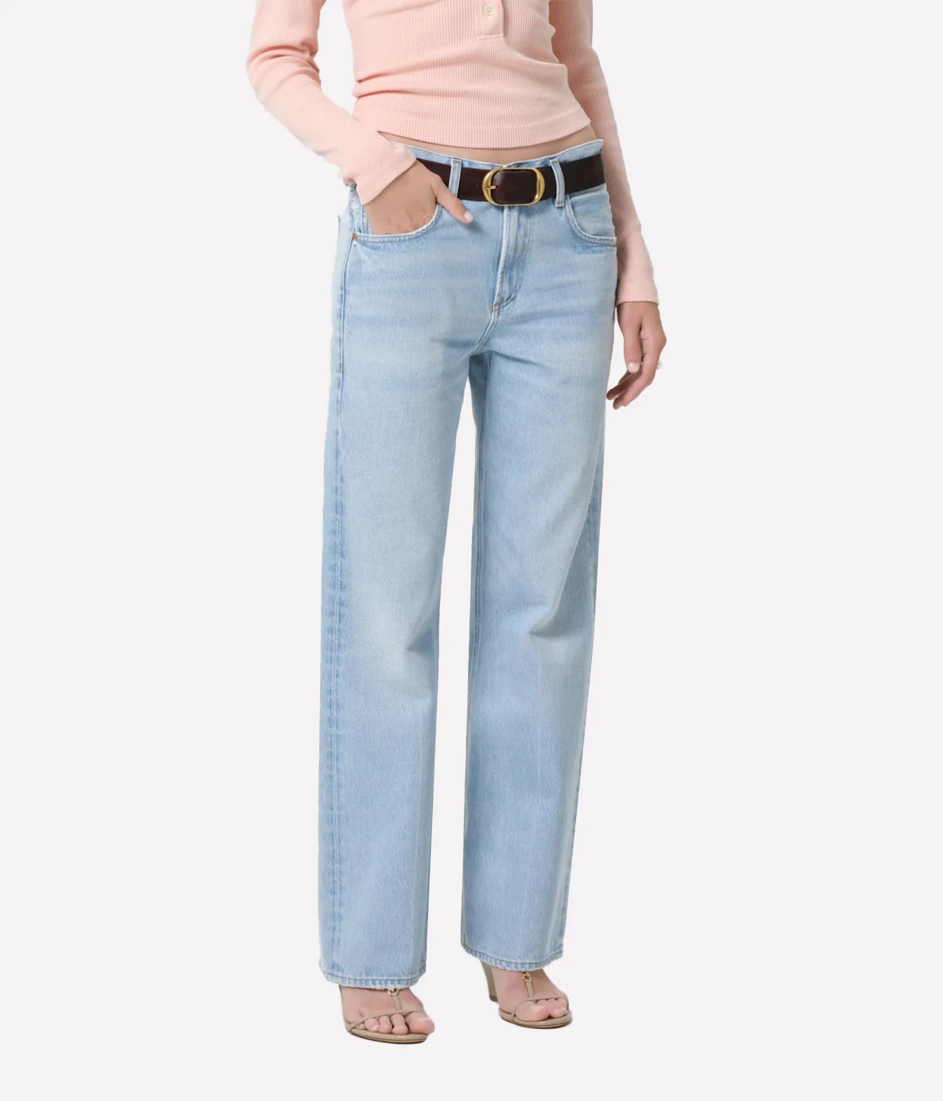 A model wearing light-wash blue straight-leg jeans with a black belt, styled with a peach-pink long-sleeve top and beige sandals, set against a plain light gray background.