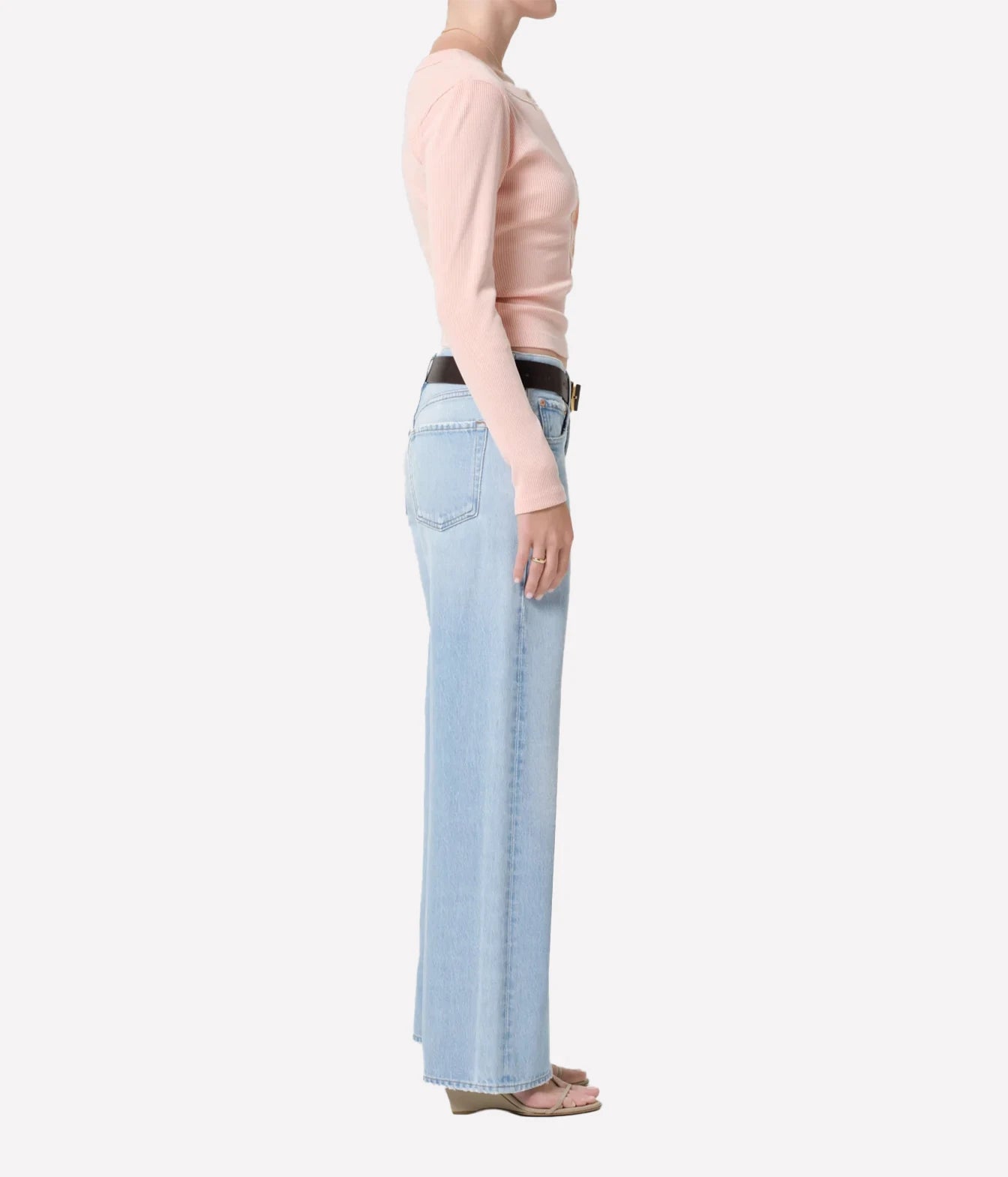A side-profile view of a model wearing light-wash wide-leg jeans, a pale pink long-sleeve top, and a black belt against a plain light gray background.