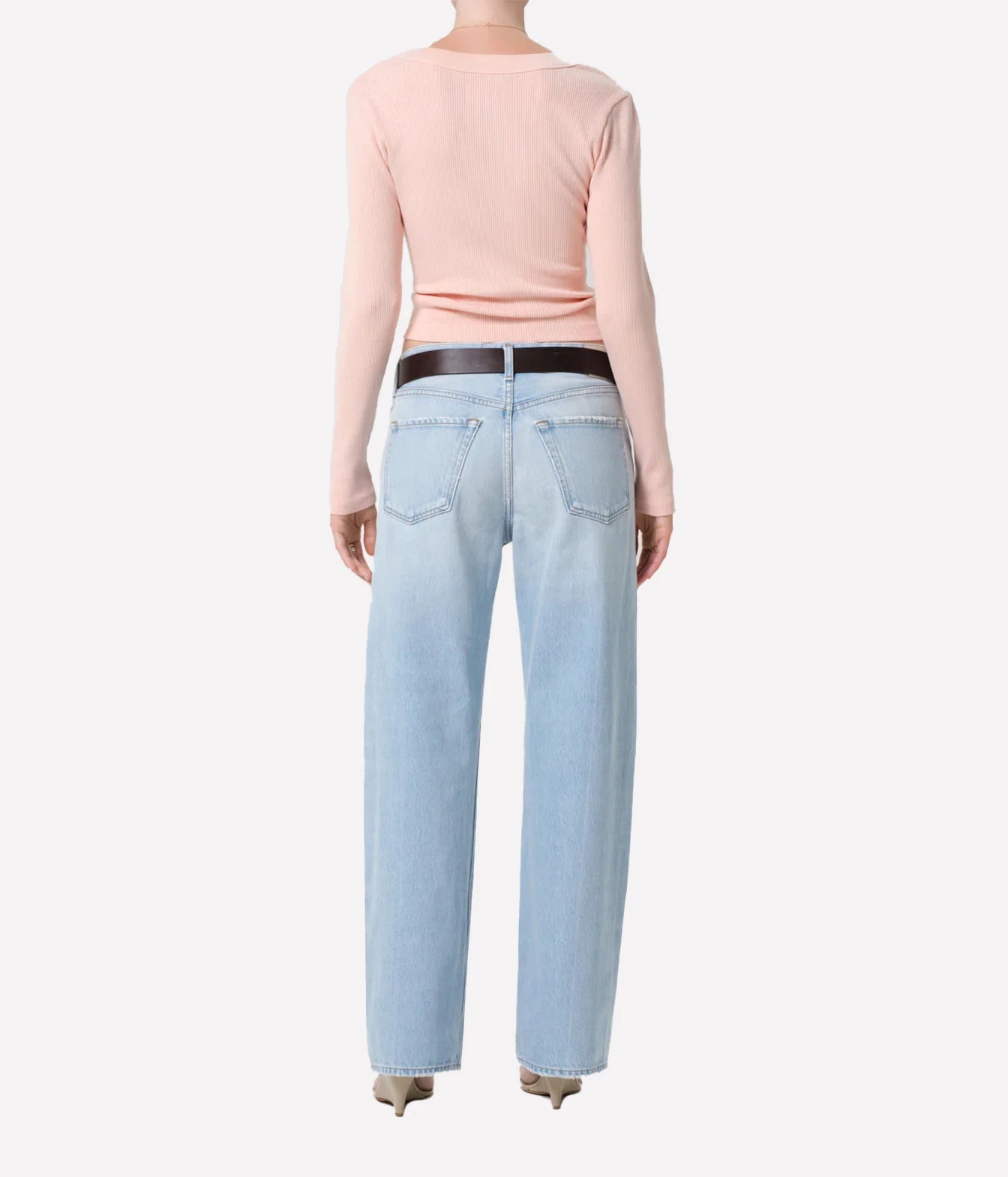 A model shown from the back wearing a light pink long-sleeve top, light-wash blue jeans with a black belt, and beige pointed-toe heels against a plain white background.