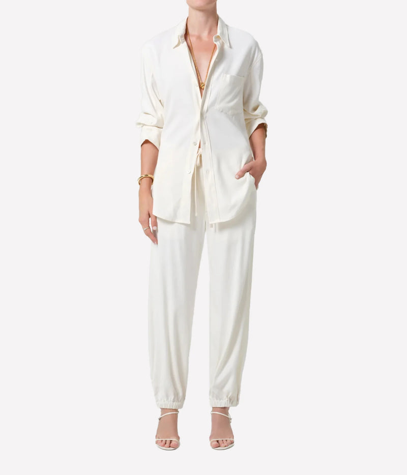 A model wearing a matching cream-colored two-piece lounge set consisting of a long-sleeve button-down shirt and coordinating jogger pants, styled with neutral sandals and gold jewelry against a plain white background.