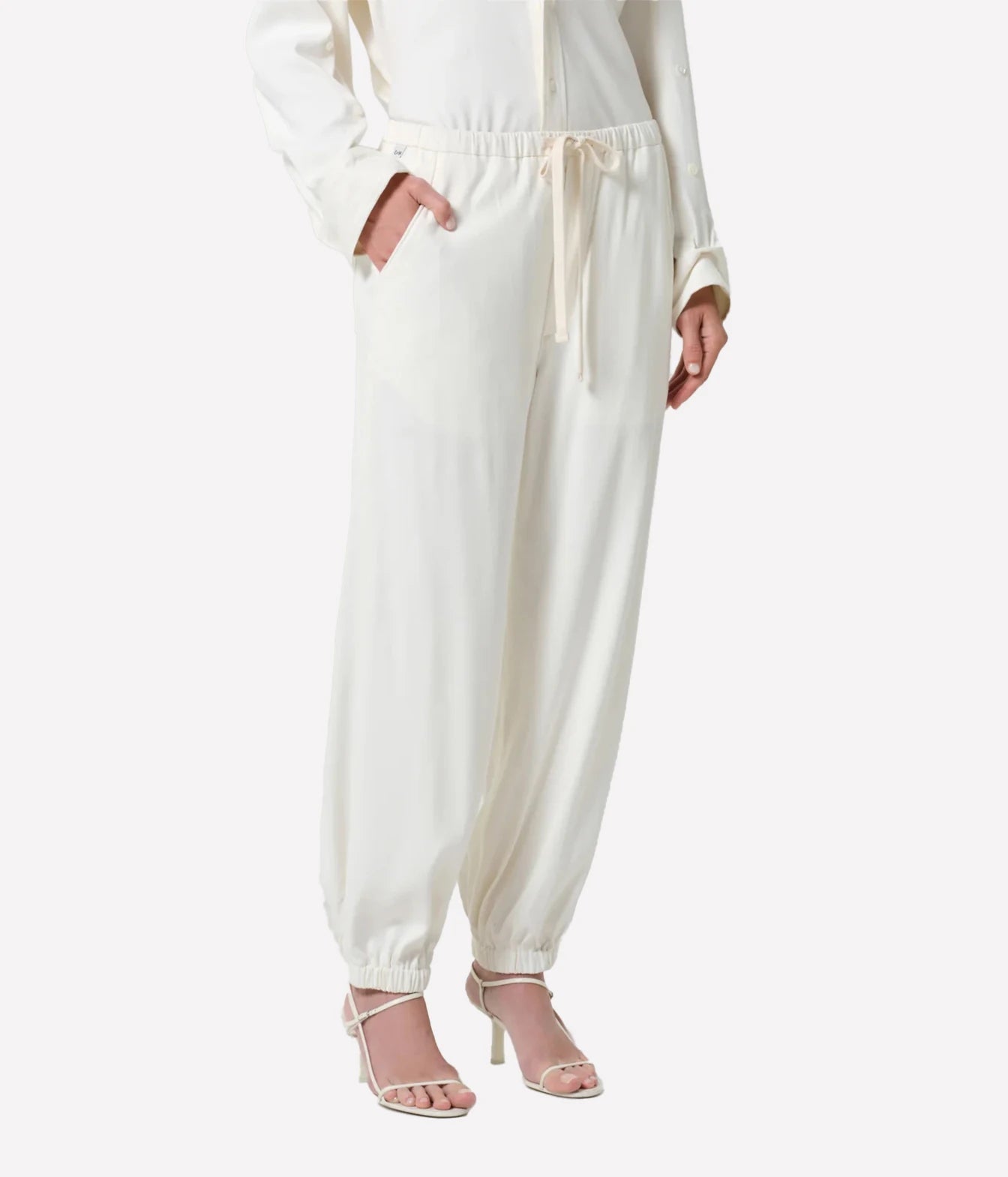 A model wearing cream-colored, wide-leg jogger pants with a drawstring waist and elastic cuffs, paired with clear high-heeled sandals against a light gray background.