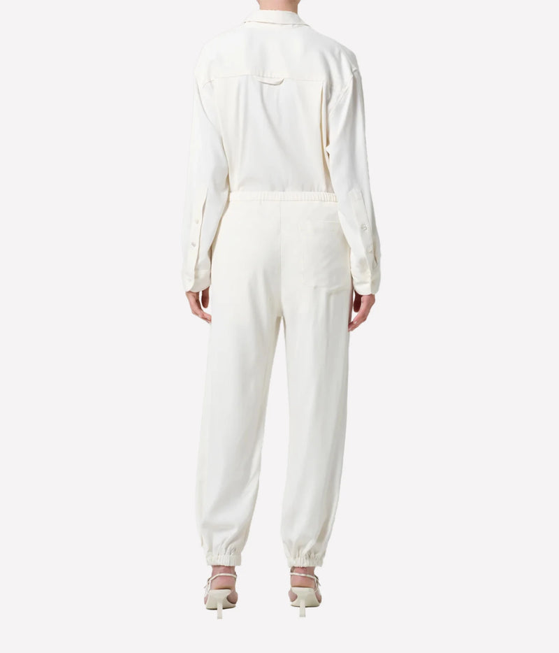 A model viewed from the back wearing a cream-colored long-sleeve jumpsuit with a relaxed fit and matching cream high-heeled sandals, set against a plain light gray studio background.