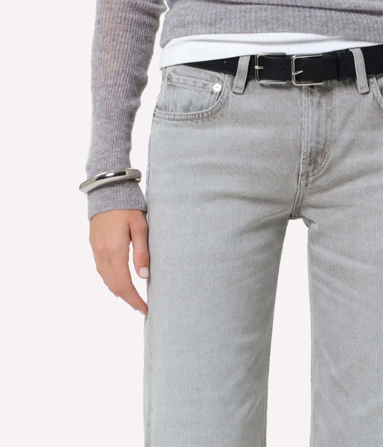 Close-up of a person wearing light gray denim jeans with a black belt, a gray ribbed sweater, and a white undershirt, against a plain light gray background.