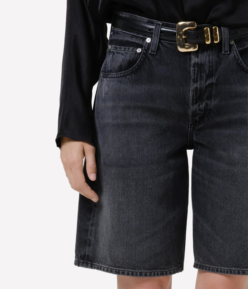 Close-up of the Citizens of Humanity Court Denim Short in Vintage Black showing the faded black wash, front pockets and fly front, worn with a black leather belt with a chunky gold buckle and gold keepers and a black satin top