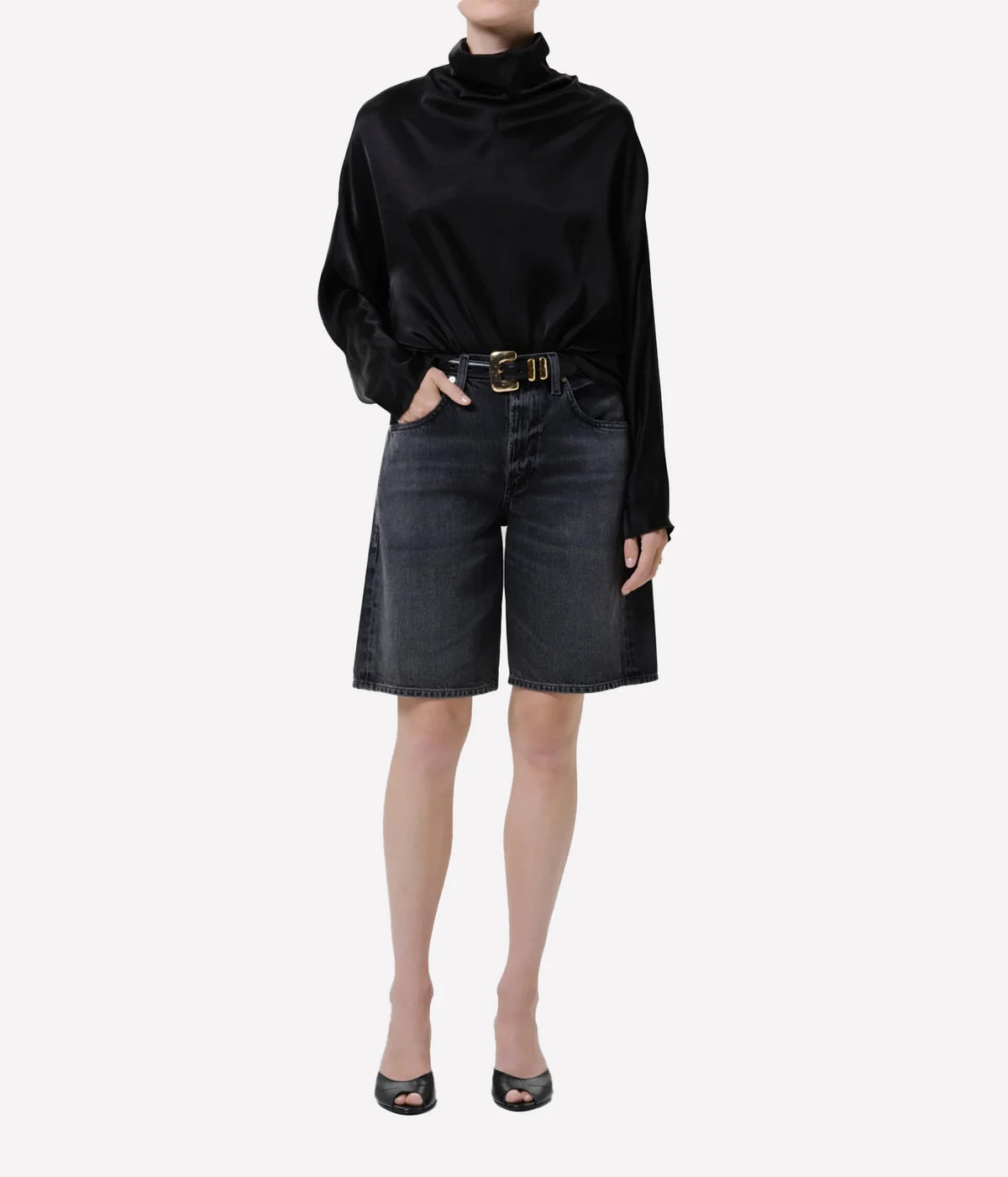 Model wearing the Citizens of Humanity Court Denim Short in Vintage Black with a black satin high-neck top, a black leather belt with a gold buckle and black open-toe kitten heel mules, one hand in her pocket