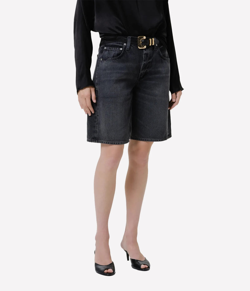 Front view of a model wearing the Citizens of Humanity Court Denim Short in Vintage Black with a tucked-in black satin top, a black leather belt with a gold buckle and black open-toe kitten heel mules, showing the relaxed wide leg and above-the-knee length