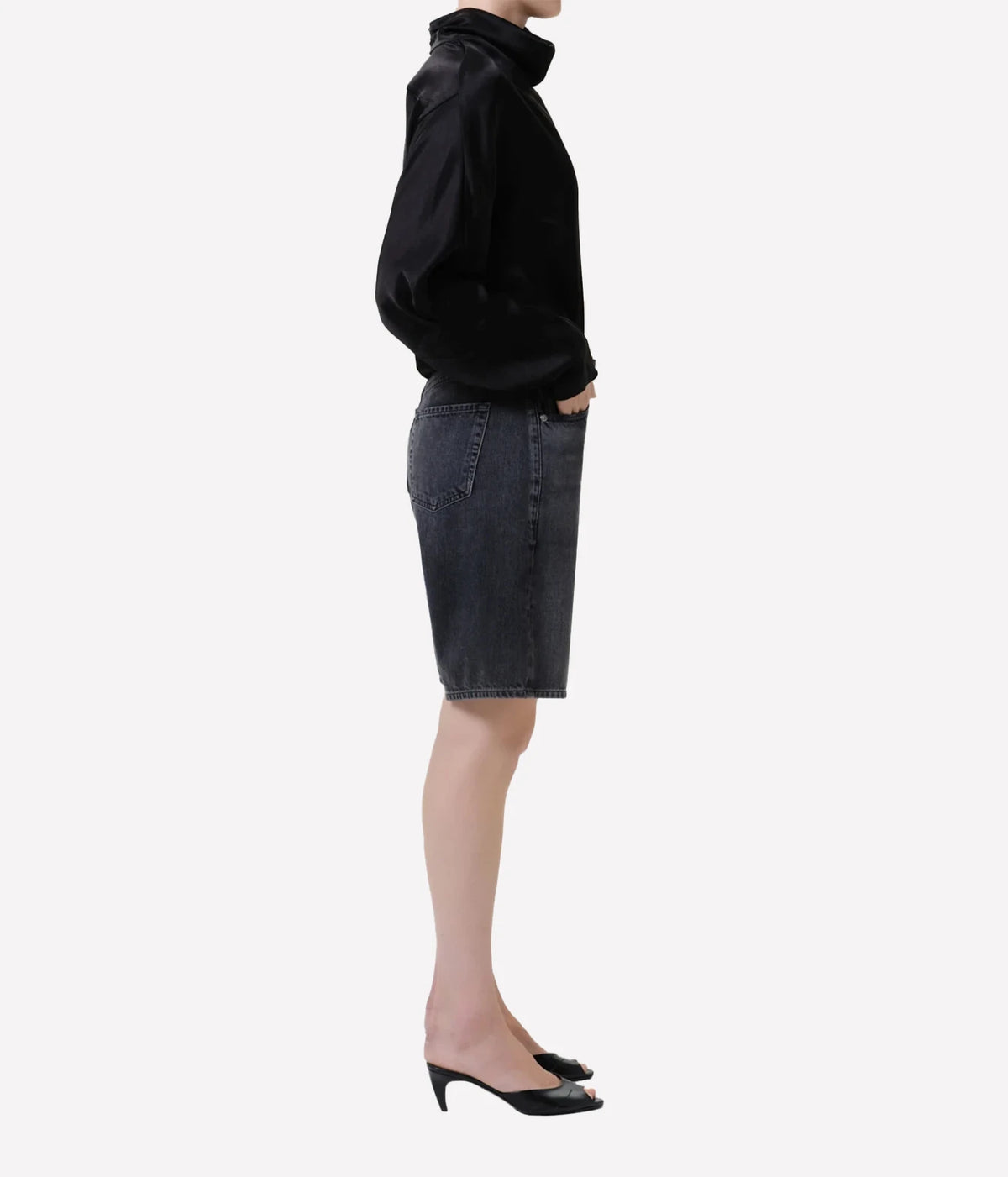 Side profile of a model wearing the Citizens of Humanity Court Denim Short in Vintage Black with a black satin high-neck top and black open-toe kitten heel mules, showing the relaxed fit and above-the-knee length