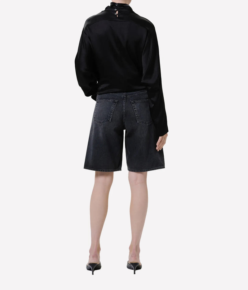 Back view of a model wearing the Citizens of Humanity Court Denim Short in Vintage Black with a black satin high-neck top and black kitten heel mules, showing the back patch pockets and wide leg