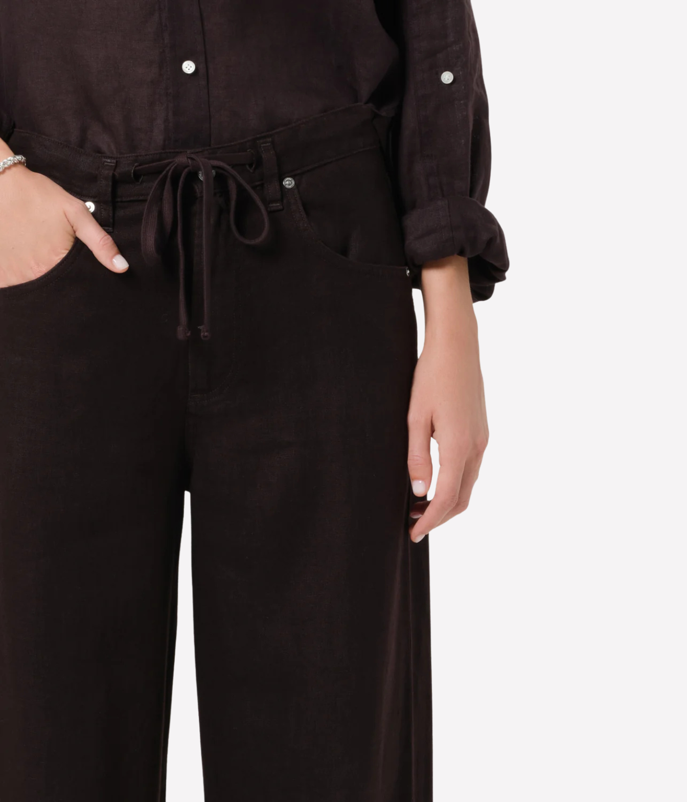 A close-up of a model wearing dark brown wide-leg trousers with a matching drawstring belt and a dark brown button-up shirt, shown against a plain light gray background.