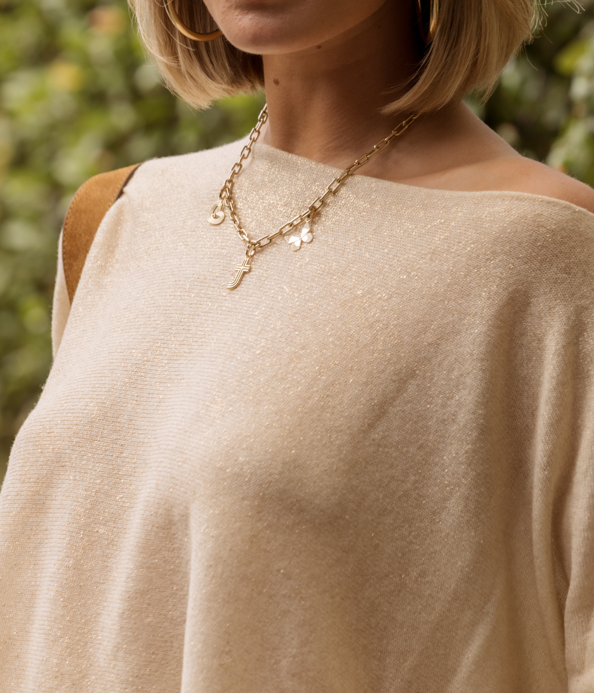 Person wearing a beige sweater and gold necklace with greenery in the background