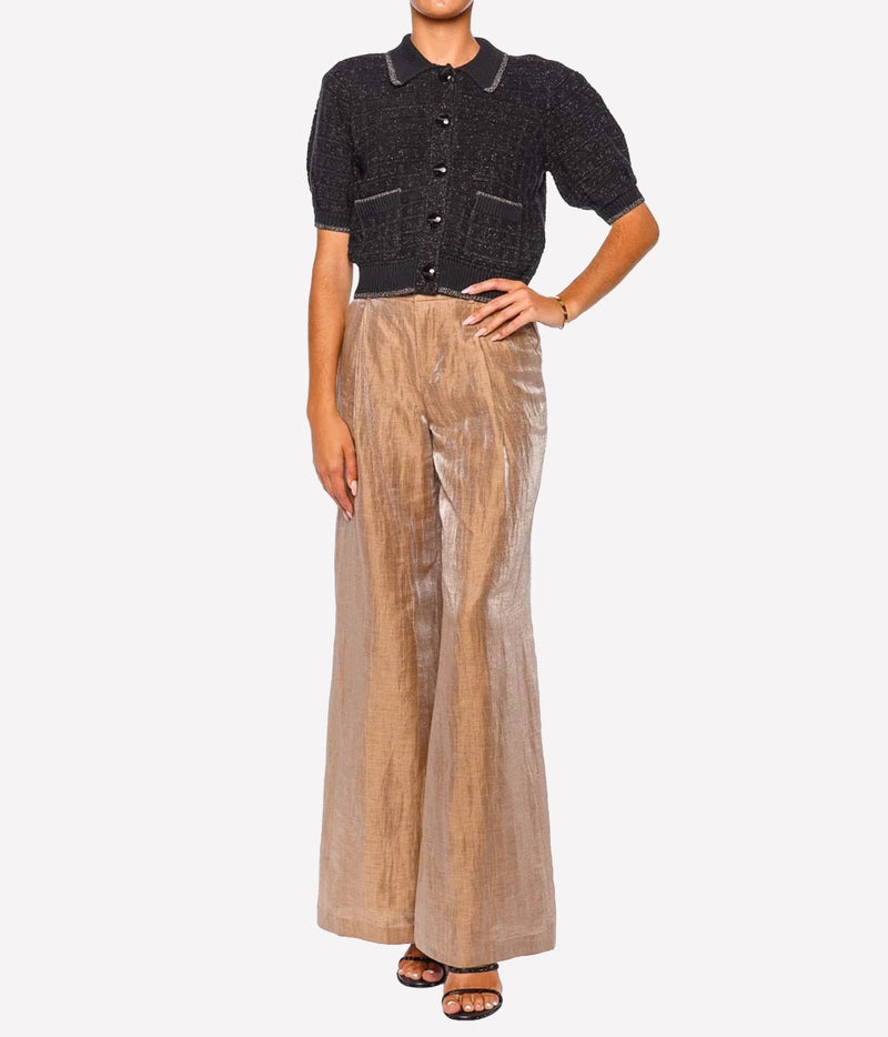 A model wearing a short-sleeve black knit button-up top with pockets, paired with high-waisted, wide-leg metallic champagne trousers, on a light gray background.
