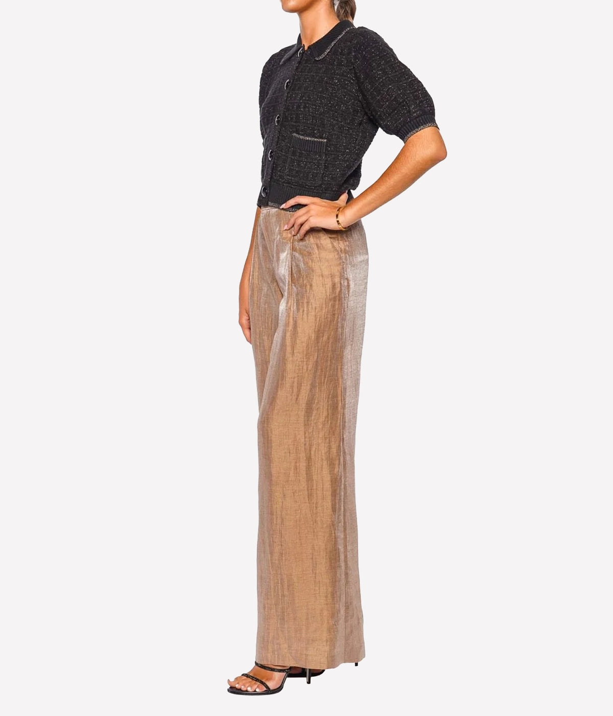 A model shown from the neck down wears a black short-sleeve knit top and high-waisted, wide-leg beige trousers with a subtle metallic sheen, paired with black heeled sandals.
