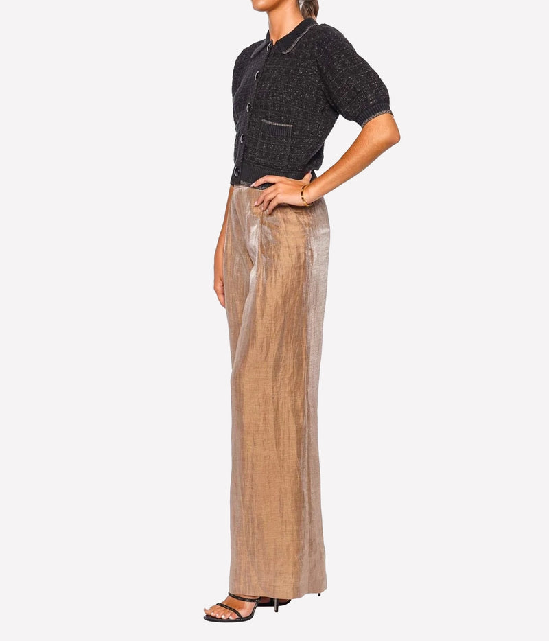 A model shown from the neck down wears a black short-sleeve knit top and high-waisted, wide-leg beige trousers with a subtle metallic sheen, paired with black heeled sandals.