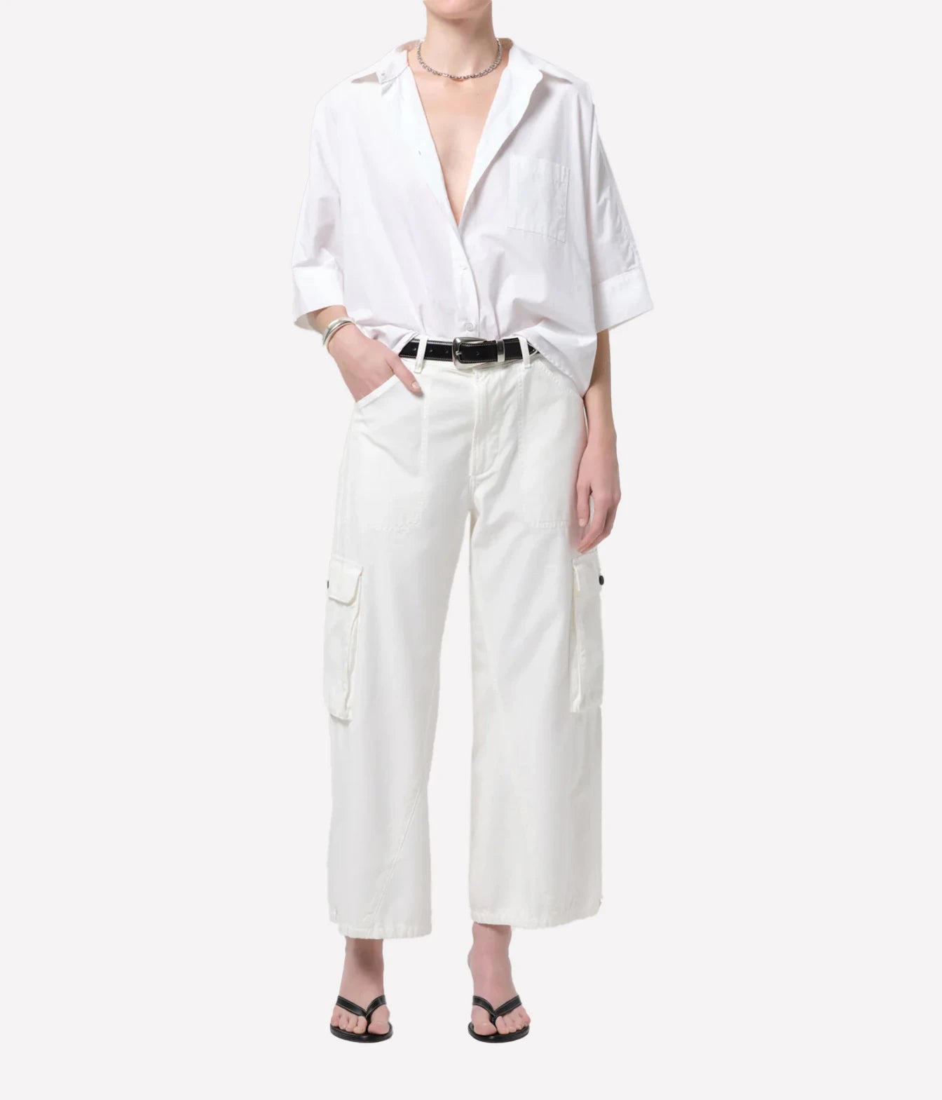 A model wearing a white short-sleeve button-up shirt and white wide-leg cargo pants with a black belt, styled with black flip-flop sandals against a light gray background.