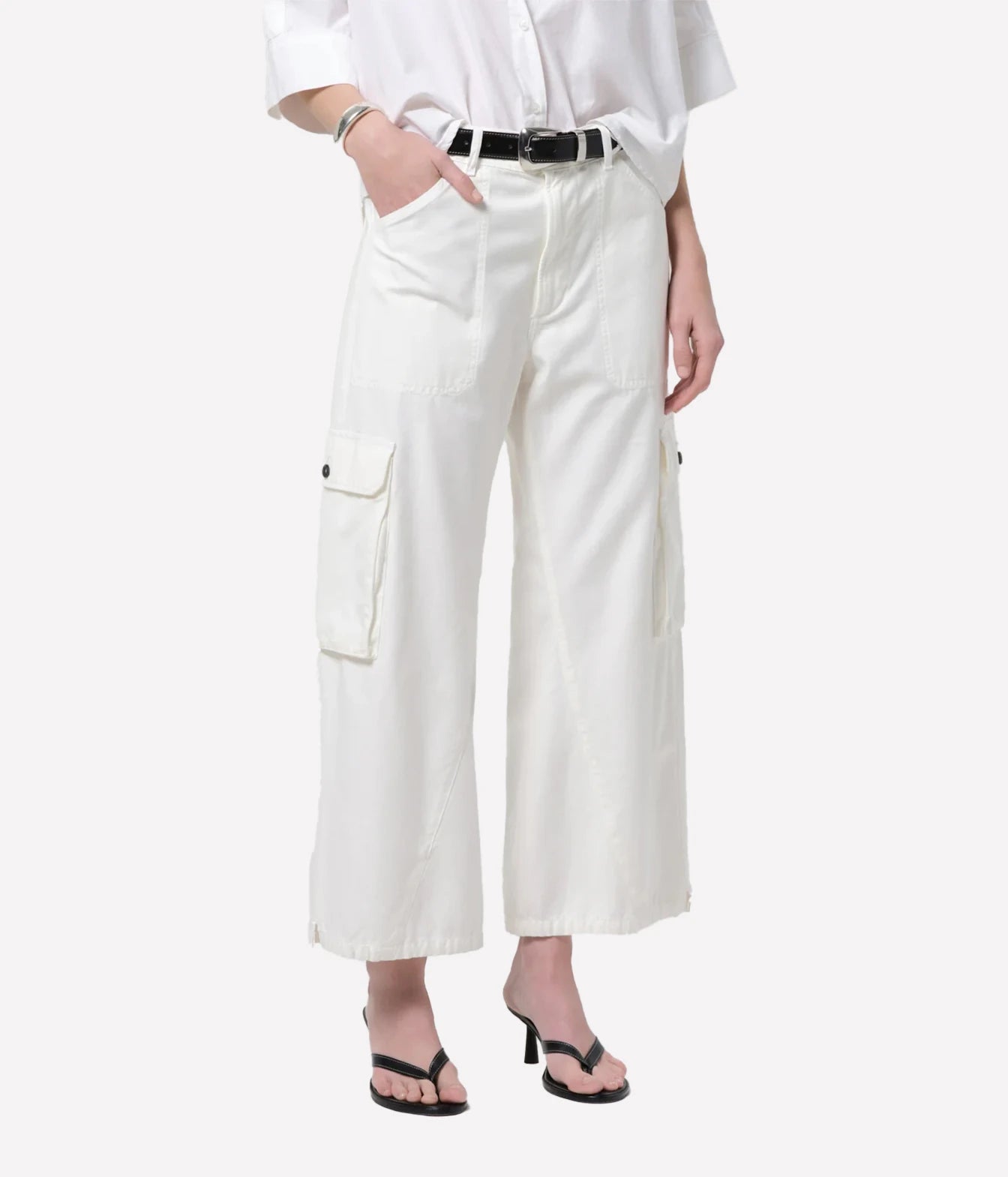 A model wearing off-white cropped cargo pants with side flap pockets, styled with a black belt, a white shirt, and black sandals against a light gray background.