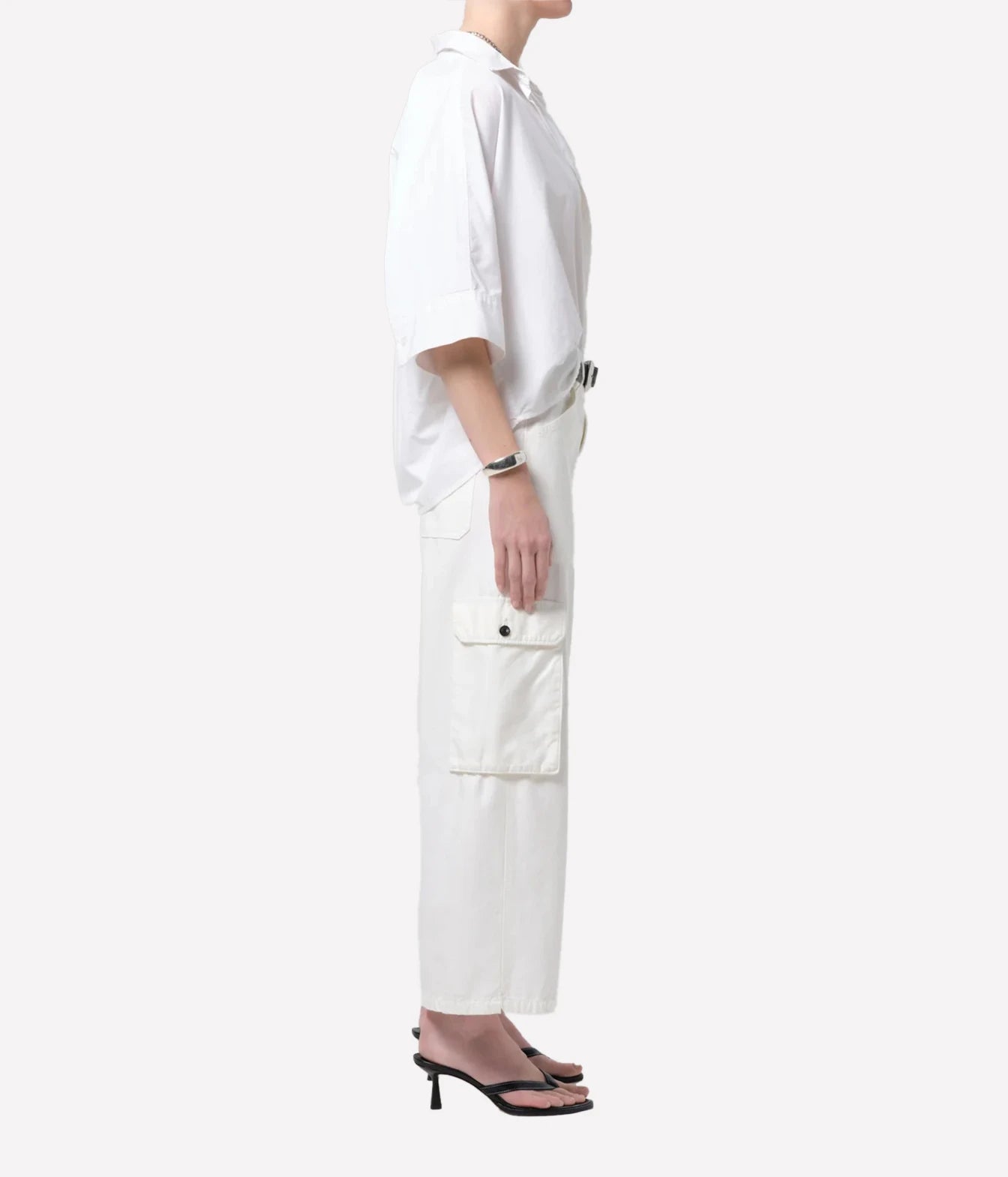 A side-profile view of a model wearing a white short-sleeve button-up shirt and matching white cargo pants with a black belt, paired with black heeled sandals, against a solid off-white background.