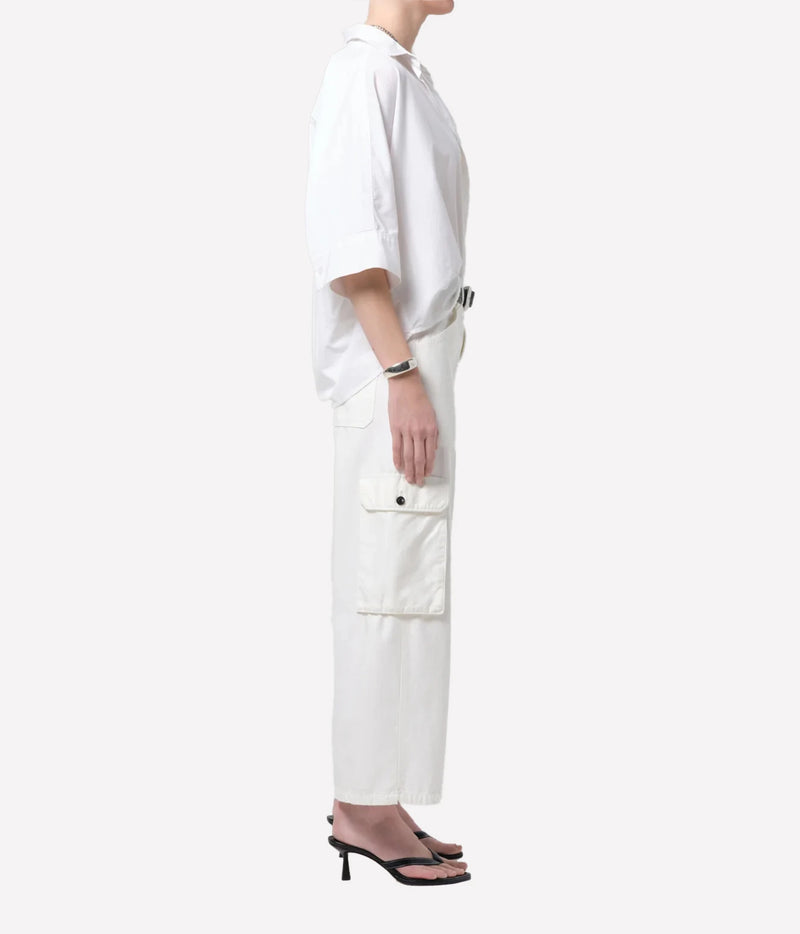 A side-profile view of a model wearing a white short-sleeve button-up shirt and matching white cargo pants with a black belt, paired with black heeled sandals, against a solid off-white background.