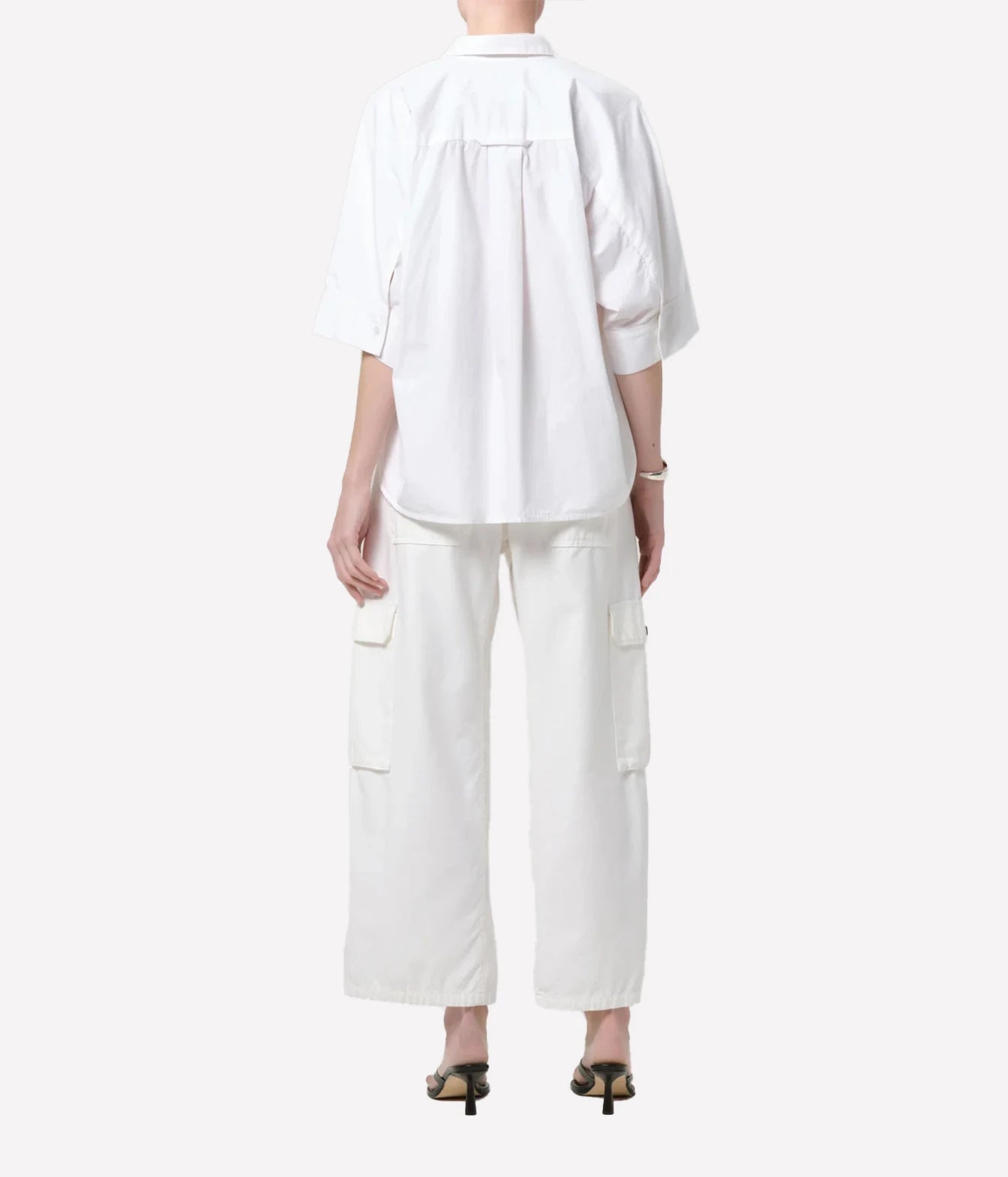 A model shown from the back wearing a white short-sleeve button-up shirt and matching white cargo pants, paired with black heeled sandals, against a light gray studio background.