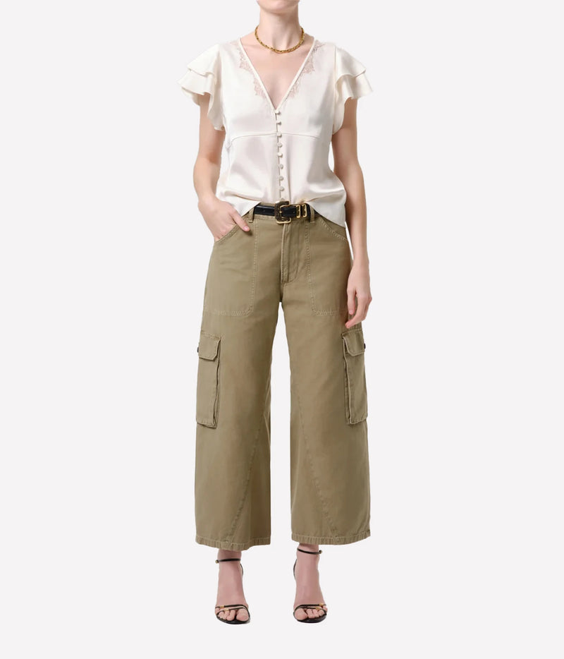 A model wearing a cream-colored short-sleeve blouse with ruffled shoulders and buttons down the front, paired with wide-leg olive cargo pants and black strappy heels, against a plain light gray background.