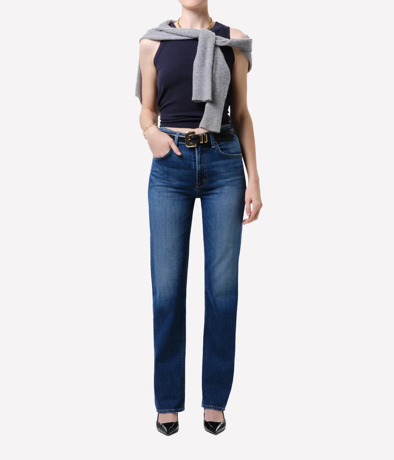 A model wearing a pair of dark blue straight-leg jeans with a gold-tone belt, styled with a navy sleeveless top, a gray knit wrap, and black pointed-toe heels against a plain white background.