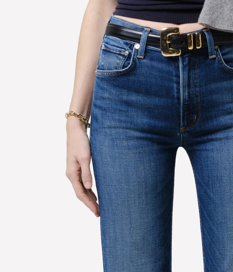 Close-up of a person wearing blue denim jeans with a black leather belt featuring a large gold-tone buckle, paired with a dark top and a gold bracelet, against a plain light background.