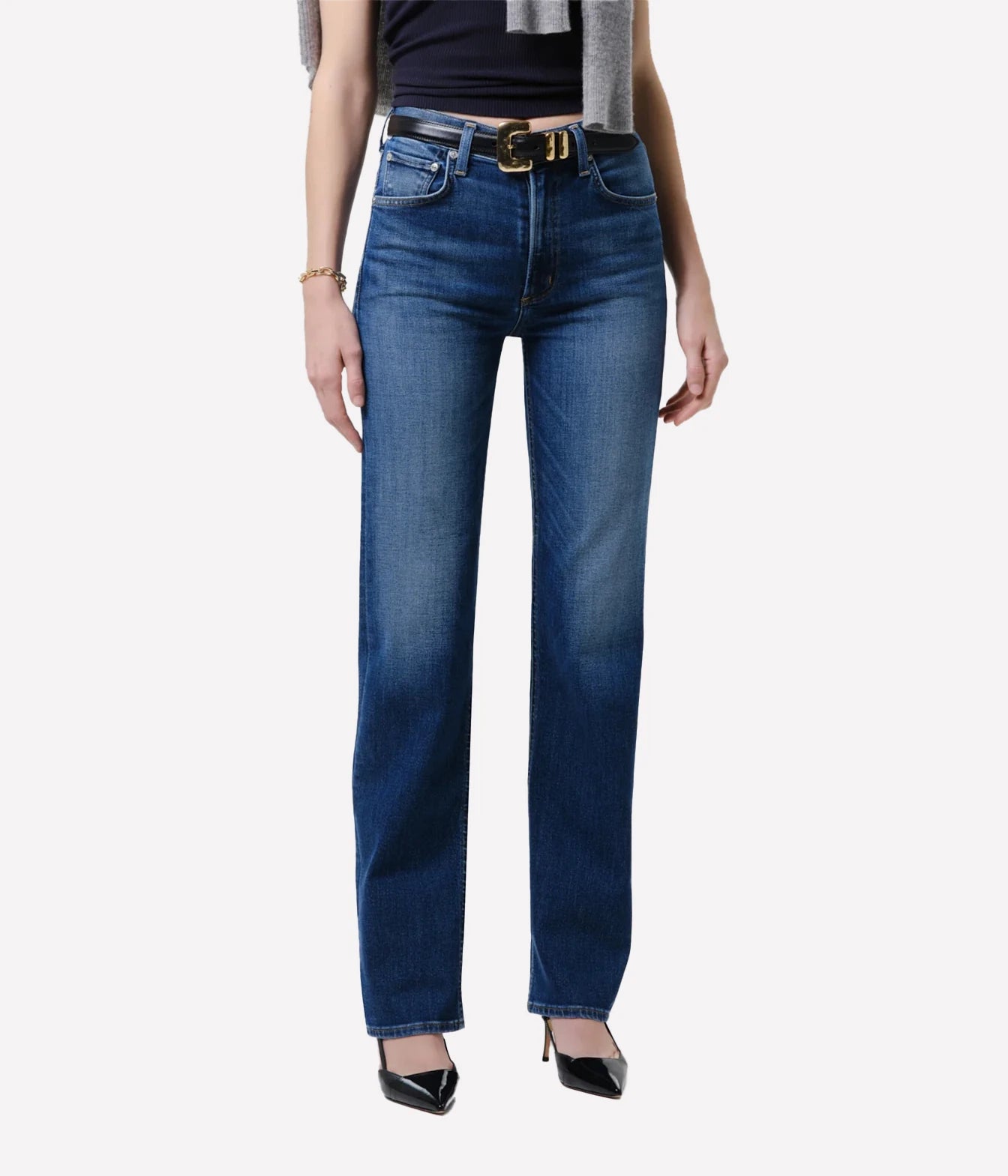 A model wearing a pair of dark blue straight-leg jeans with a black belt featuring a gold buckle, styled with a black top, gray cardigan, and black pointed-toe heels against a light gray background.
