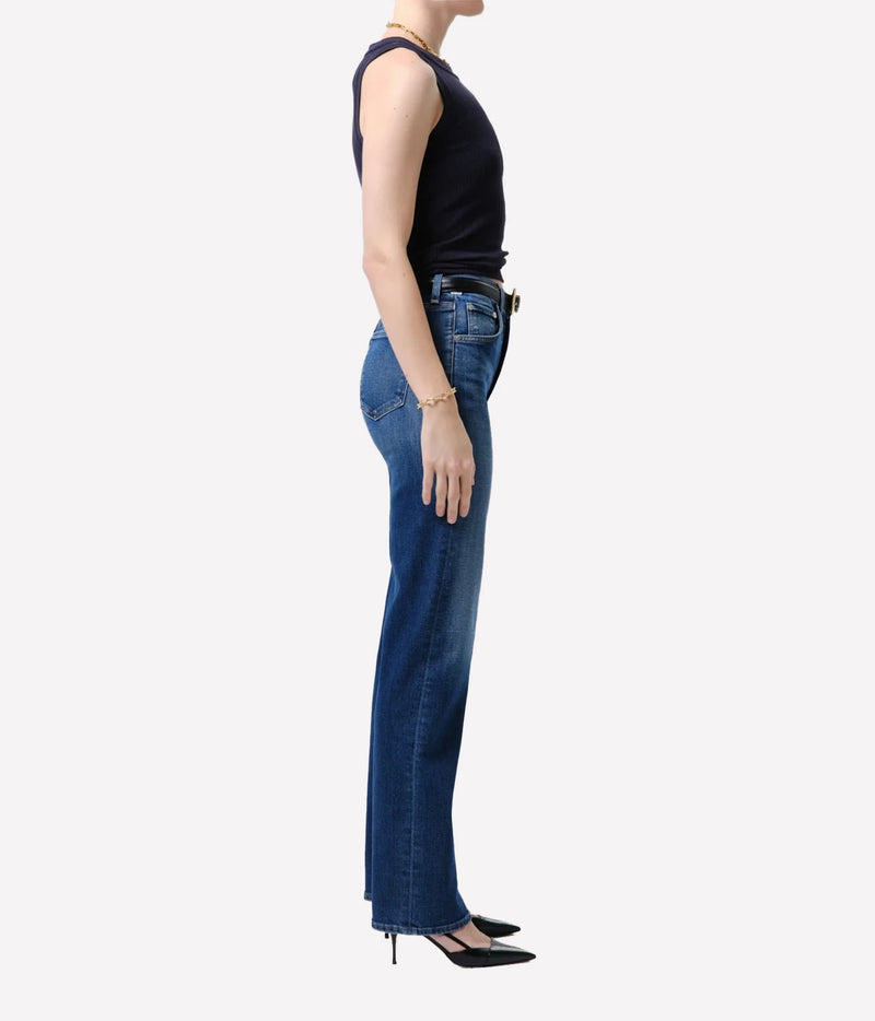 A side-profile studio shot of a model wearing high-waisted, wide-leg blue jeans, a black sleeveless top, and black heels against a light gray background.