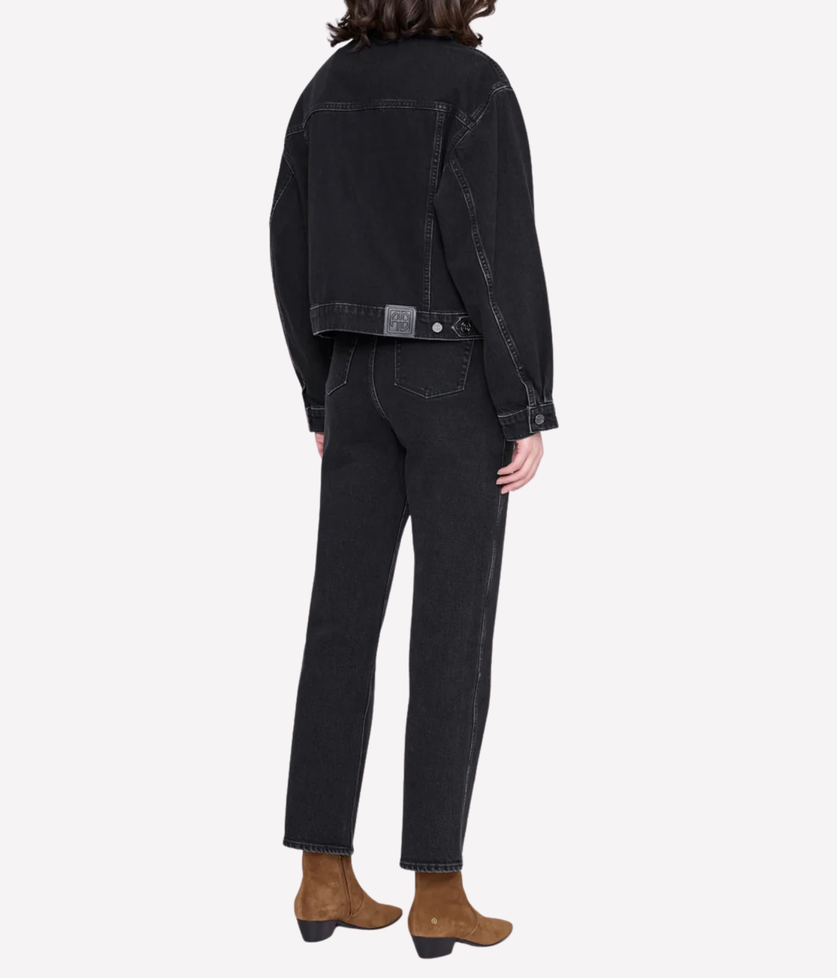 A model shown from the back wearing a black cropped denim jacket and black straight-leg jeans, paired with brown ankle boots, against a light gray studio background.