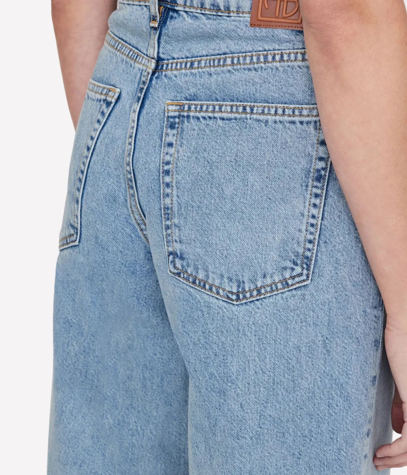 Close-up back view of a person wearing light blue denim jeans, showing the back pocket, stitching, waistband, and a brown leather brand patch.