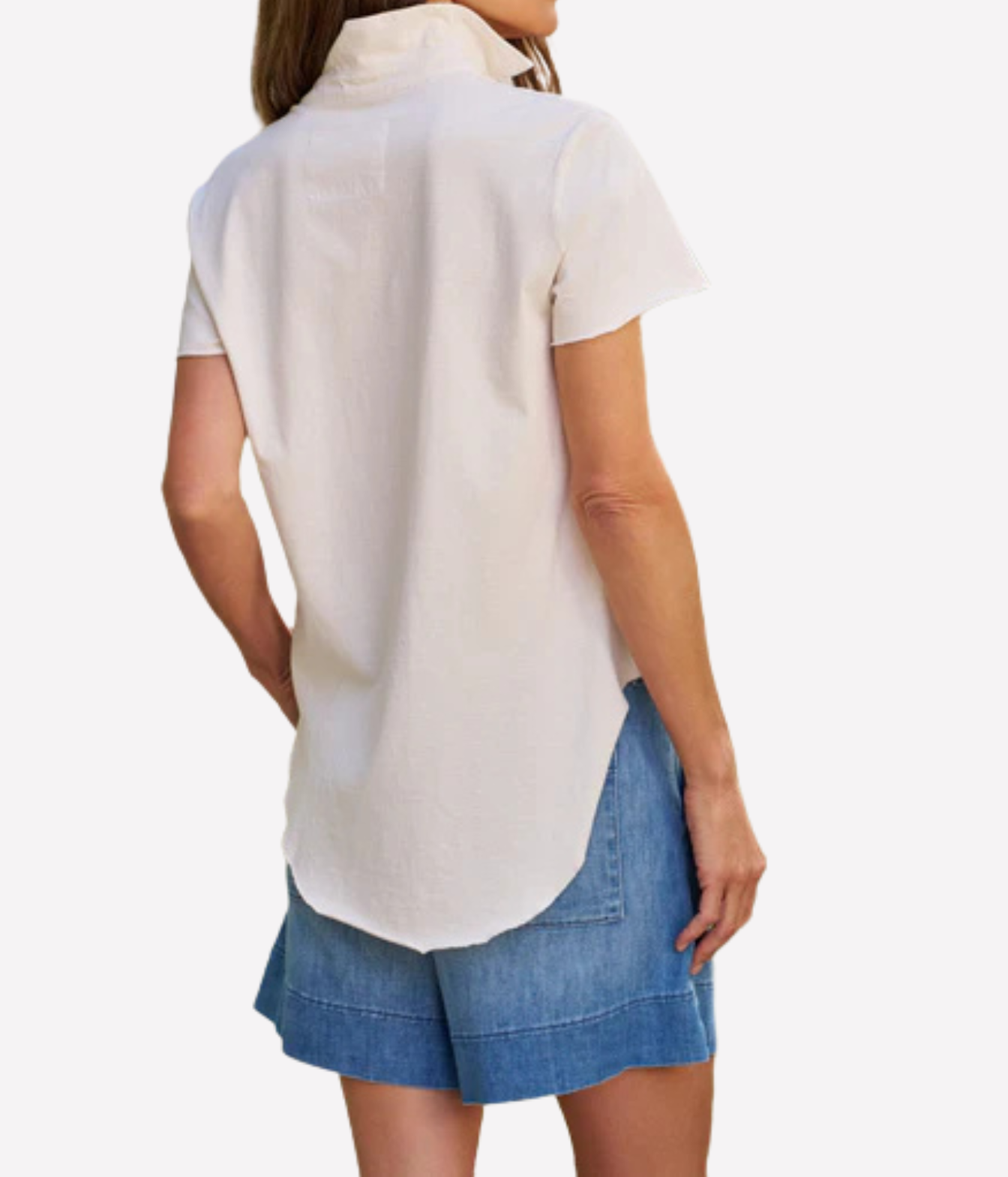 A woman shown from the back wearing a short-sleeve white button-up shirt and blue denim shorts against a plain light gray background.