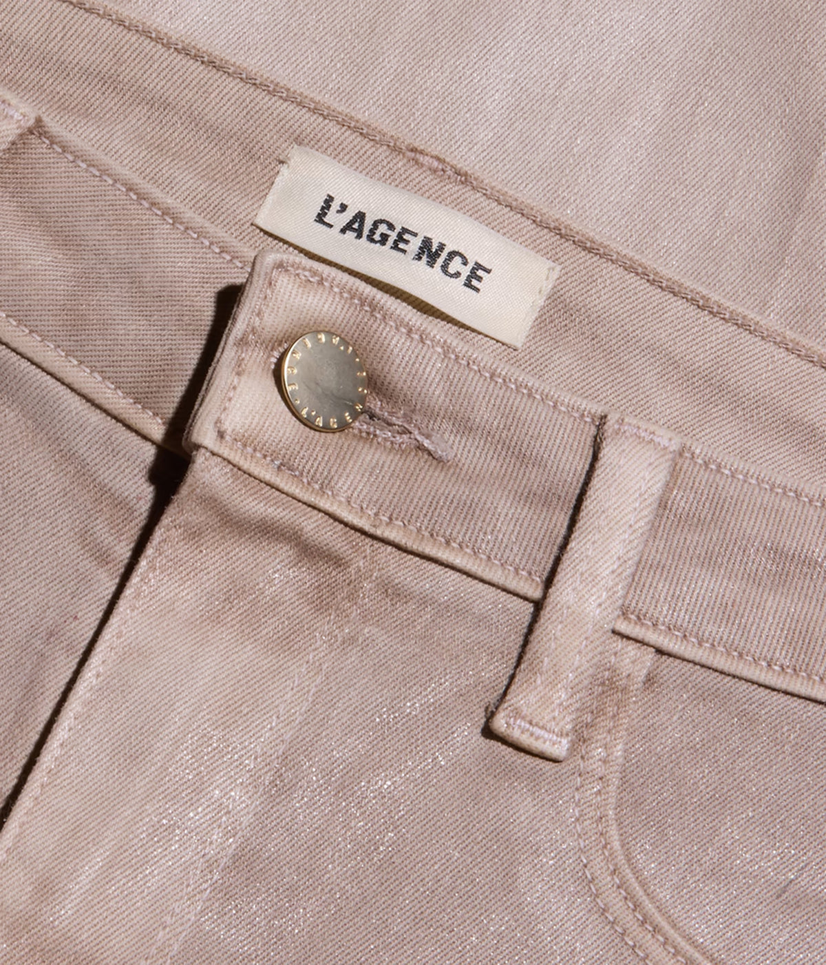 Close-up of a pair of light beige denim jeans showing the waistband, belt loop, button, and a cream-coloured fabric brand label that reads 'L'AGENCE'.