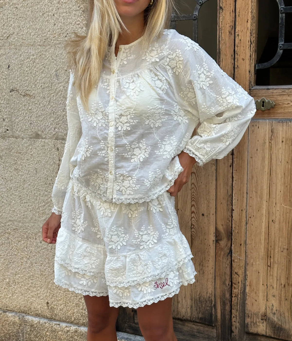 Model standing with one hand on her hip wearing the Sissel Edelbo Delphine Top in Off White buttoned with matching tiered ruffle shorts, showing the floral schiffli embroidery, gathered yoke and long gathered sleeves