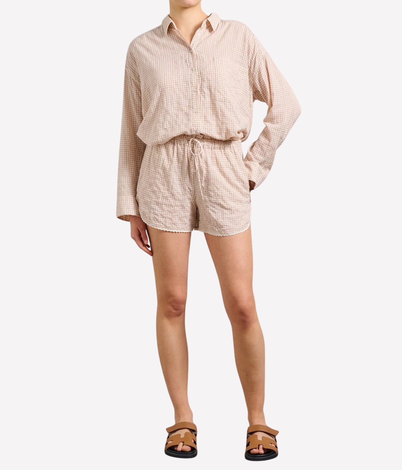 A model wearing a matching beige and white gingham two-piece set consisting of a long-sleeve button-down shirt and coordinating shorts, paired with brown sandals against a light gray background.