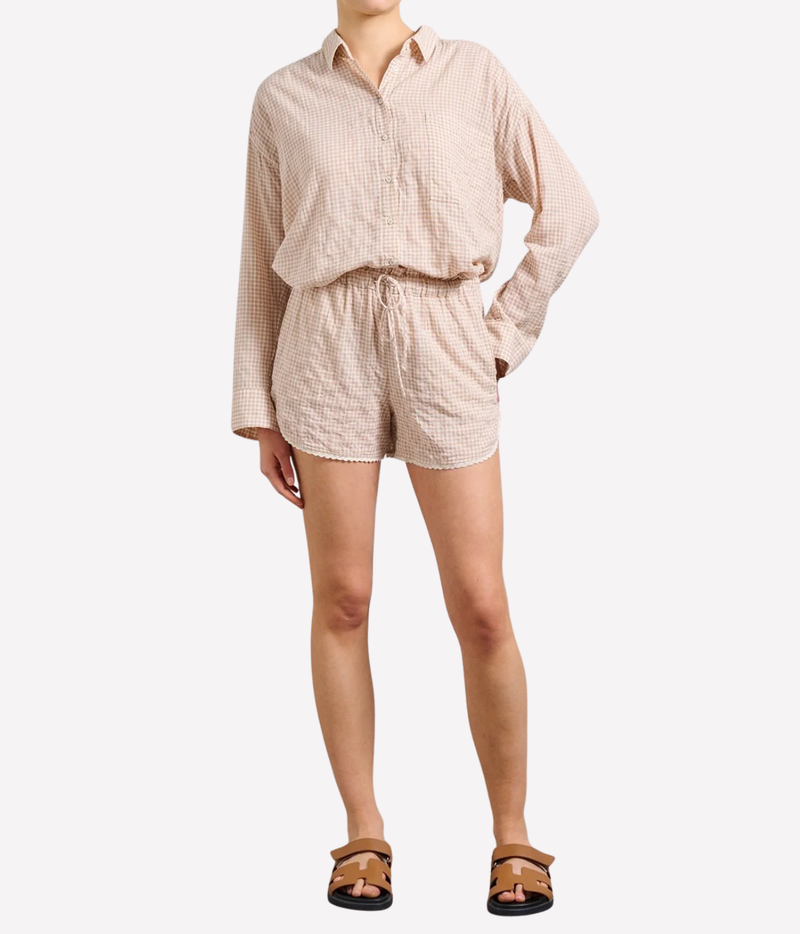 A model wearing a matching beige and white gingham two-piece set consisting of a long-sleeve button-down shirt and coordinating shorts, paired with brown sandals against a light gray background.