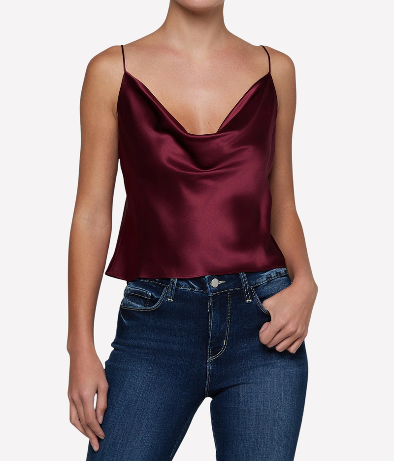A woman wearing a silk cowl-neck camisole top in deep burgundy with thin spaghetti straps, paired with dark blue jeans.