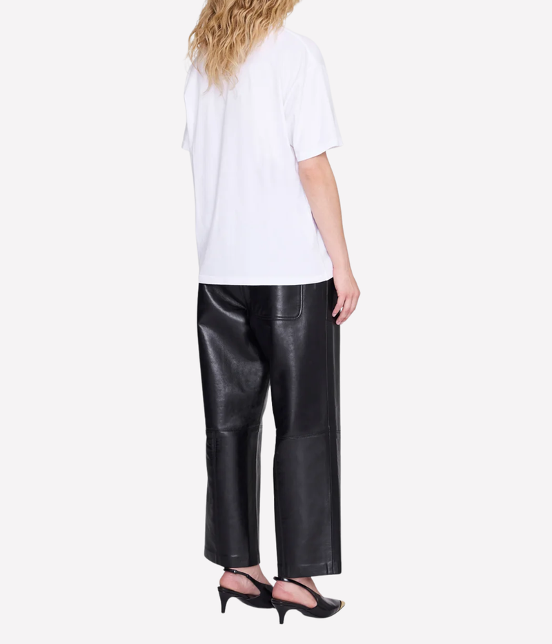 Rear three-quarter view of a model wearing the Anine Bing Cade Tee Los Angeles In White with black leather straight-leg trousers and black slingback heels, showing the dropped shoulders and relaxed boxy fit