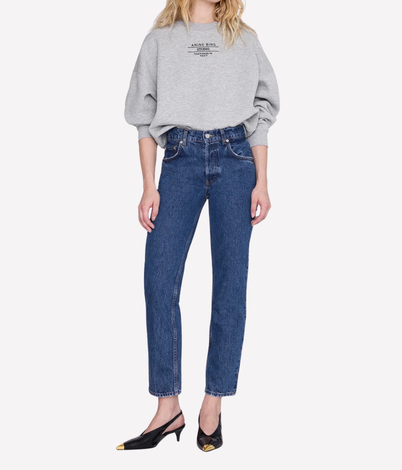 A model wearing a light gray oversized sweatshirt with black text on the chest, blue high-waisted straight-leg jeans, and black pointed-toe slingback heels with gold toe caps, set against a plain light gray background.