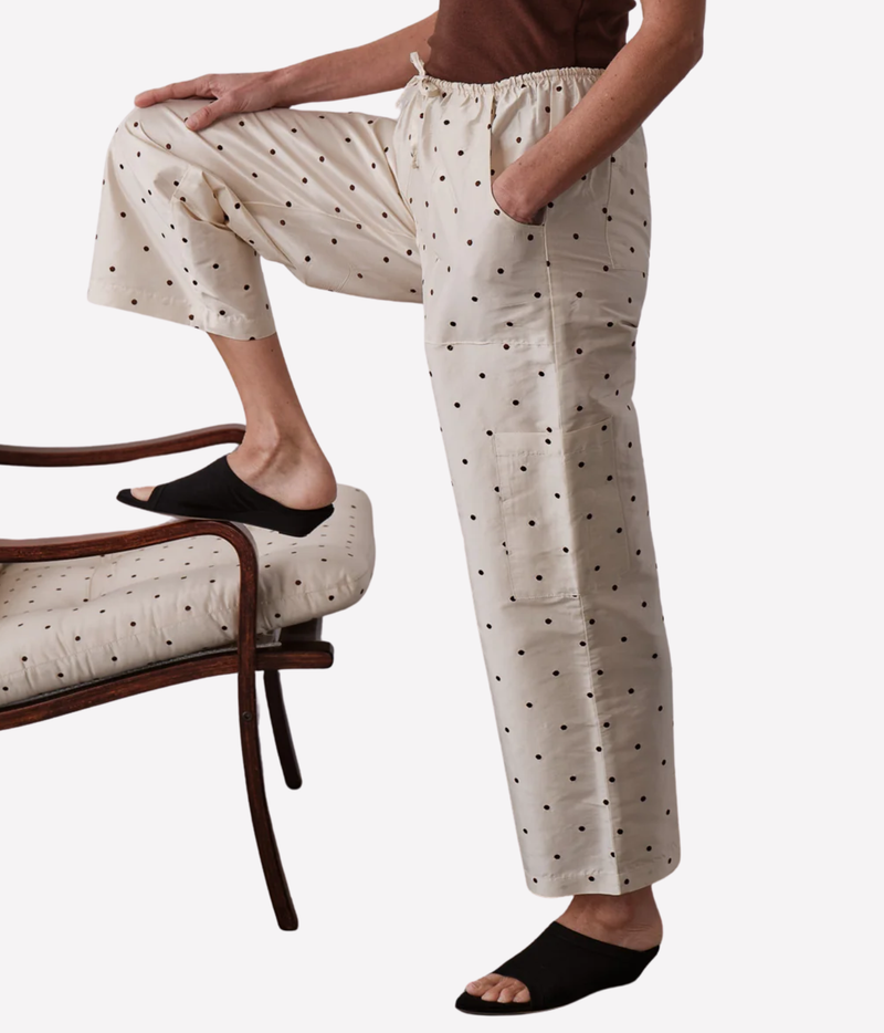 A person wearing cream-colored wide-leg pants with a black polka dot pattern, paired with black slide sandals, shown in a lifestyle pose with one leg raised on a matching cushioned chair against a light gray background.
