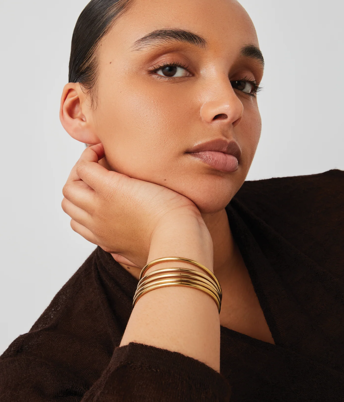 Model wearing the Jenny Bird Dane Bangle Set in Gold stacked on her wrist with her hand resting against her chin, styled with a chocolate brown top and gold earrings