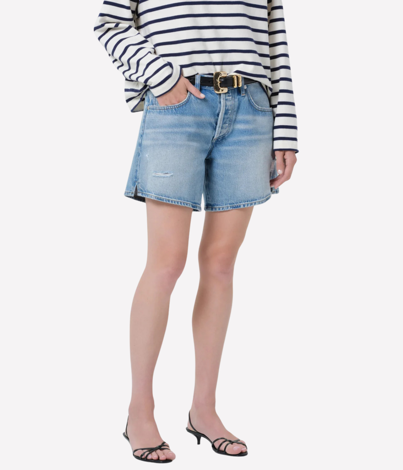 A model wearing light-wash blue denim shorts with distressed details, styled with a black and white striped long-sleeve top, a black belt with a gold buckle, and black strappy heeled sandals, set against a plain light gray background.