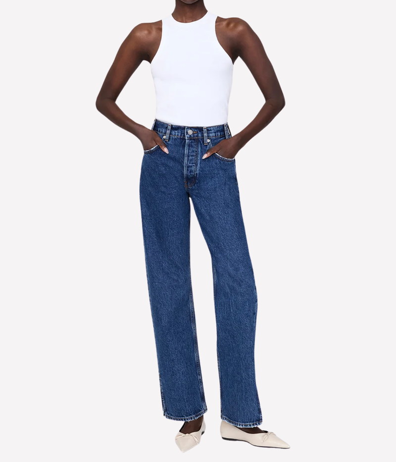 A model wearing a pair of high-waisted, wide-leg blue denim jeans with a white sleeveless top and beige pointed flats, standing against a light gray studio background.
