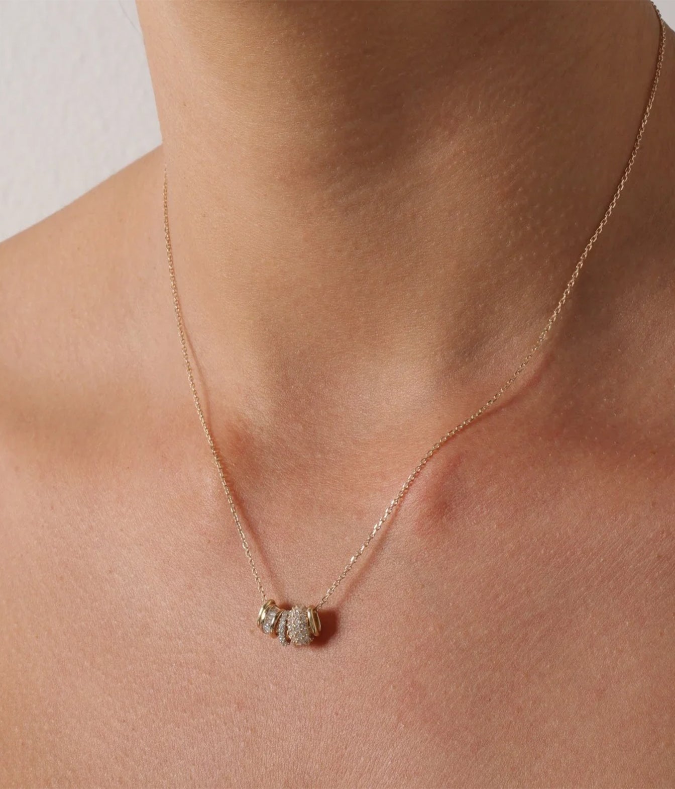 A close-up of a delicate gold chain necklace worn on a model's neck, featuring a small cylindrical pendant encrusted with clear stones.