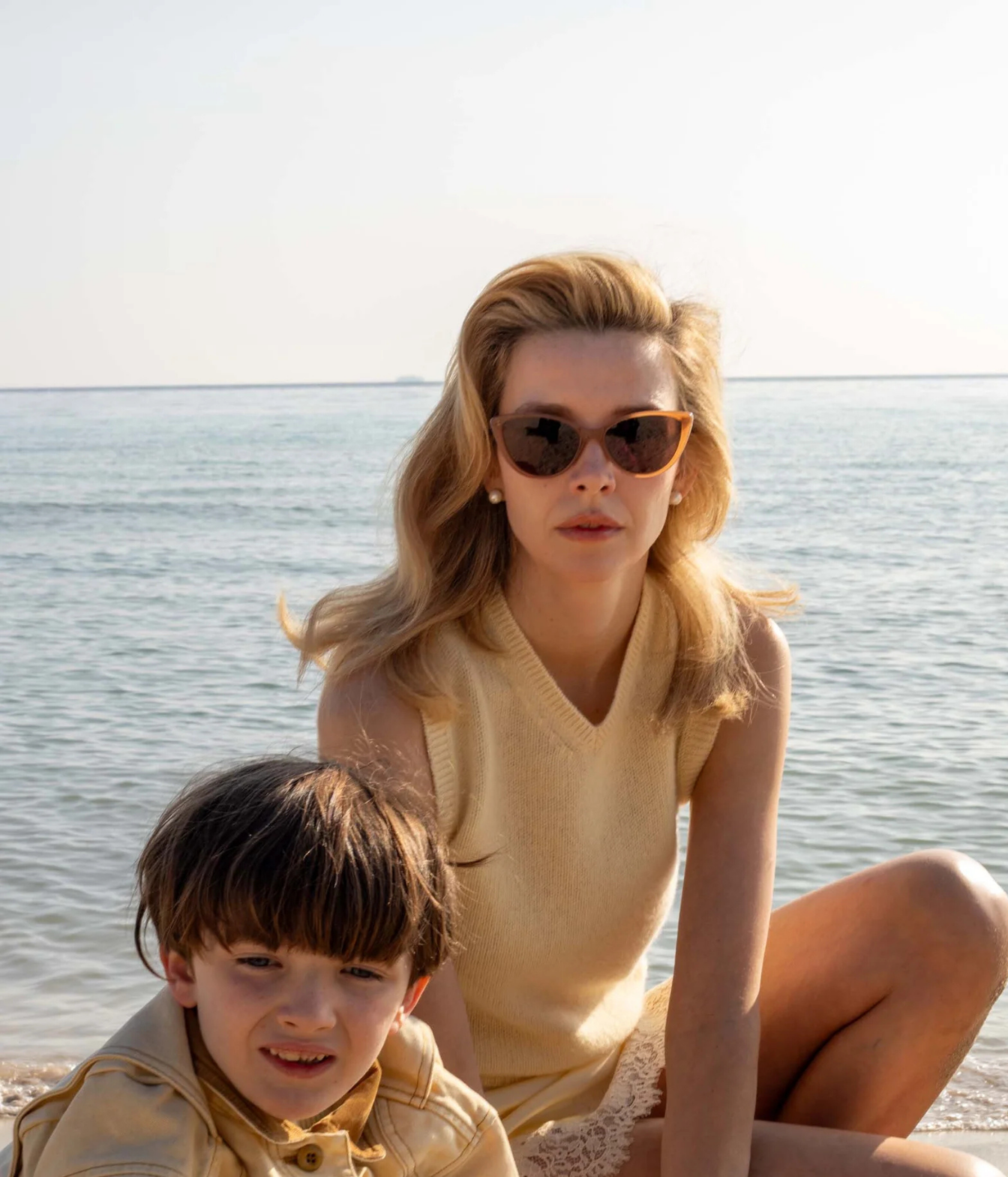 A woman and a young boy sitting on a beach shoreline, woman wearing sunglasses with brown orange frames