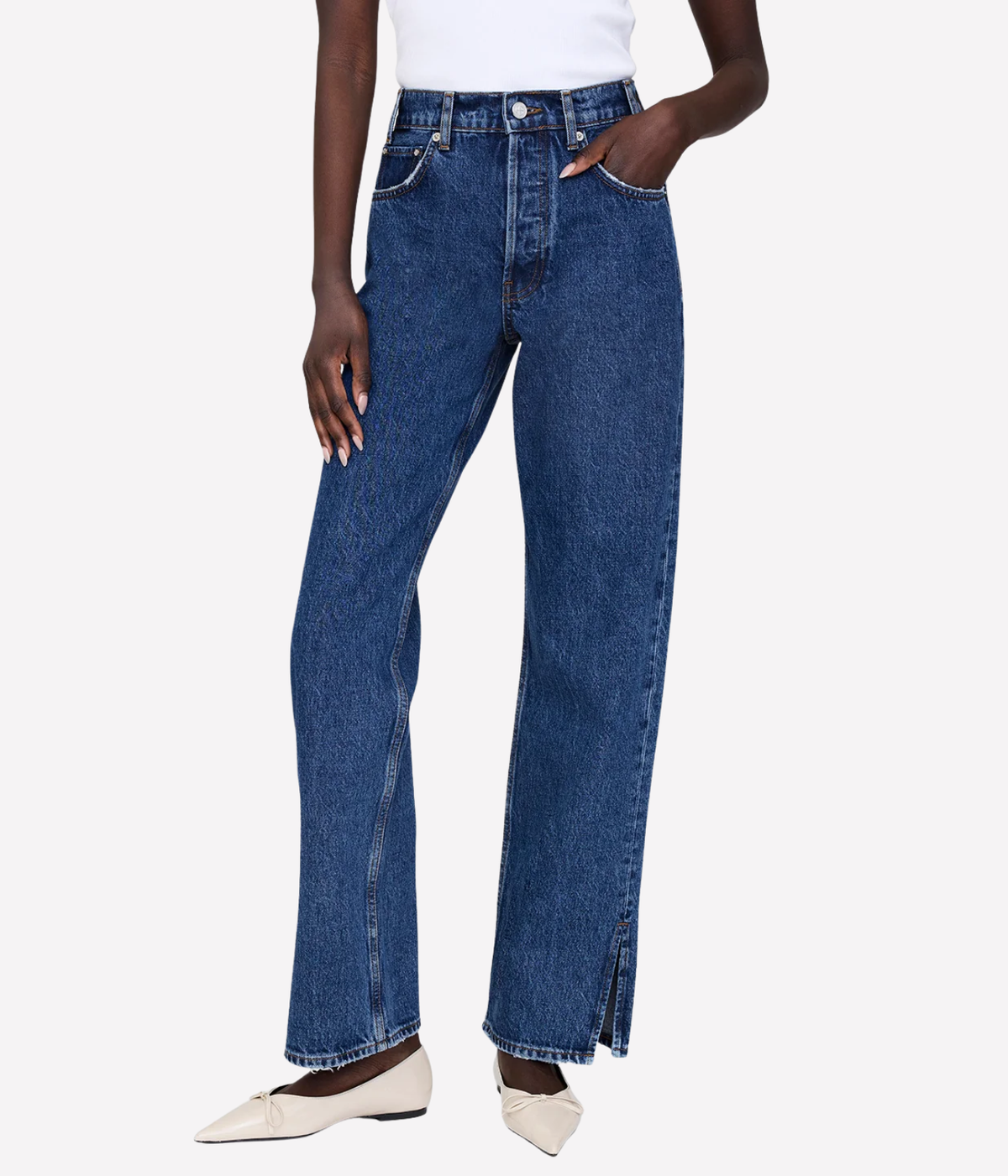 A model wearing a pair of high-waisted, wide-leg blue denim jeans with front slits at the hem, styled with a white top and beige pointed flats against a light gray background.