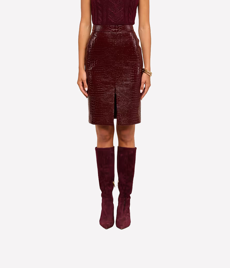 A model is shown from the chest down wearing a vegan leather, dark burgundy crocodile-embossed skirt with a front slit, paired with matching dark burgundy knee-high boots