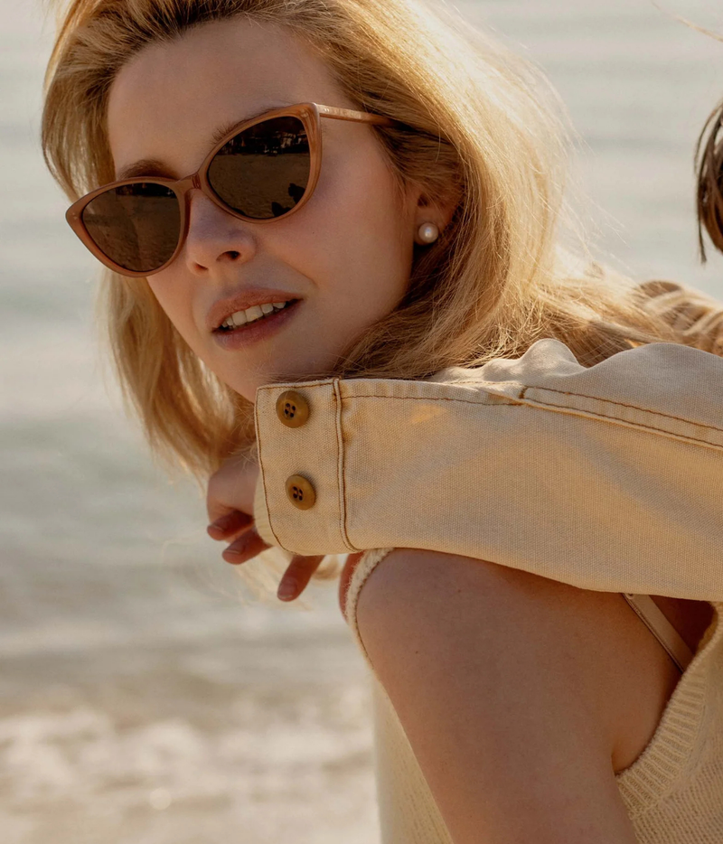 A close-up lifestyle shot of a woman wearing cat-eye sunglasses with brown frames and dark lenses
