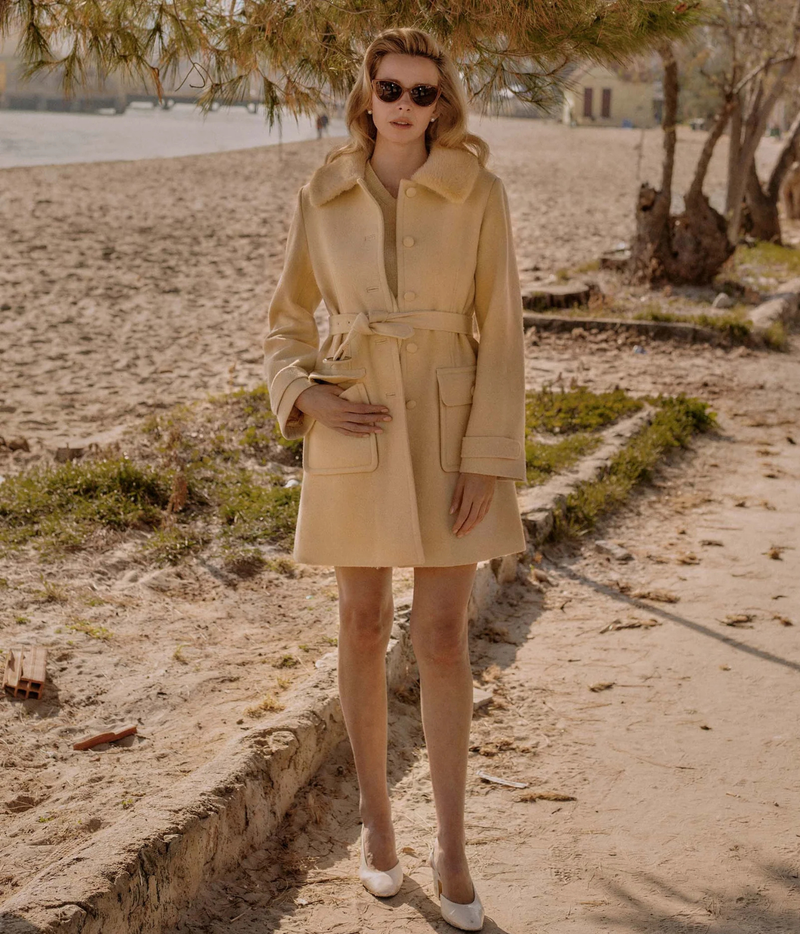 A woman stands on a sandy beach path wearing a beige belted trench coat, sunglasses