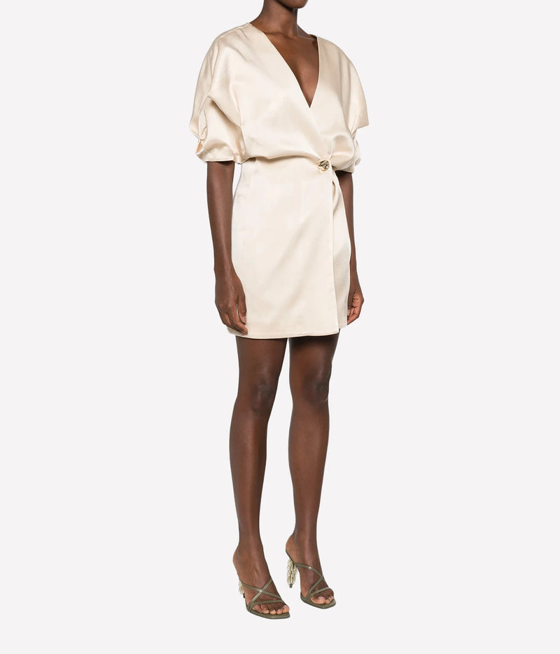 Three-quarter view of a model wearing the Raquel Diniz Anastasia Short Dress in Vanilla with strappy heeled sandals, showing the draped sleeves and wrap front