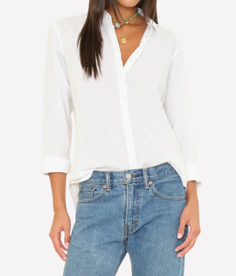 Front view of a model wearing the XiRENA Beau Shirt in White unbuttoned at the neck with cuffs rolled, loosely tucked into mid-wash straight-leg jeans and layered with a green beaded choker and gold pendant necklace